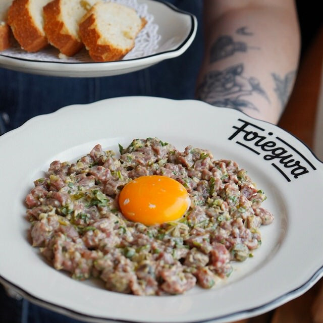 Plate of food with an egg on top, held by a person with tattoos, in front of another plate of bread.