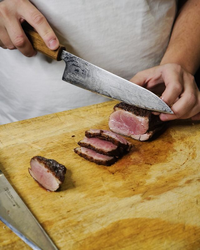 Person slicing a piece of meat on a wooden cutting board with a large knife.