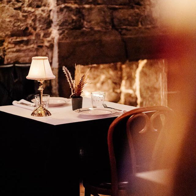 Dining table set with a lamp, candles, and decorative items against a stone wall.
