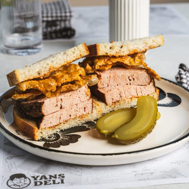 Sandwich with deli meat and pickles on a plate with 'Yans Deli' branding, placed on a table.